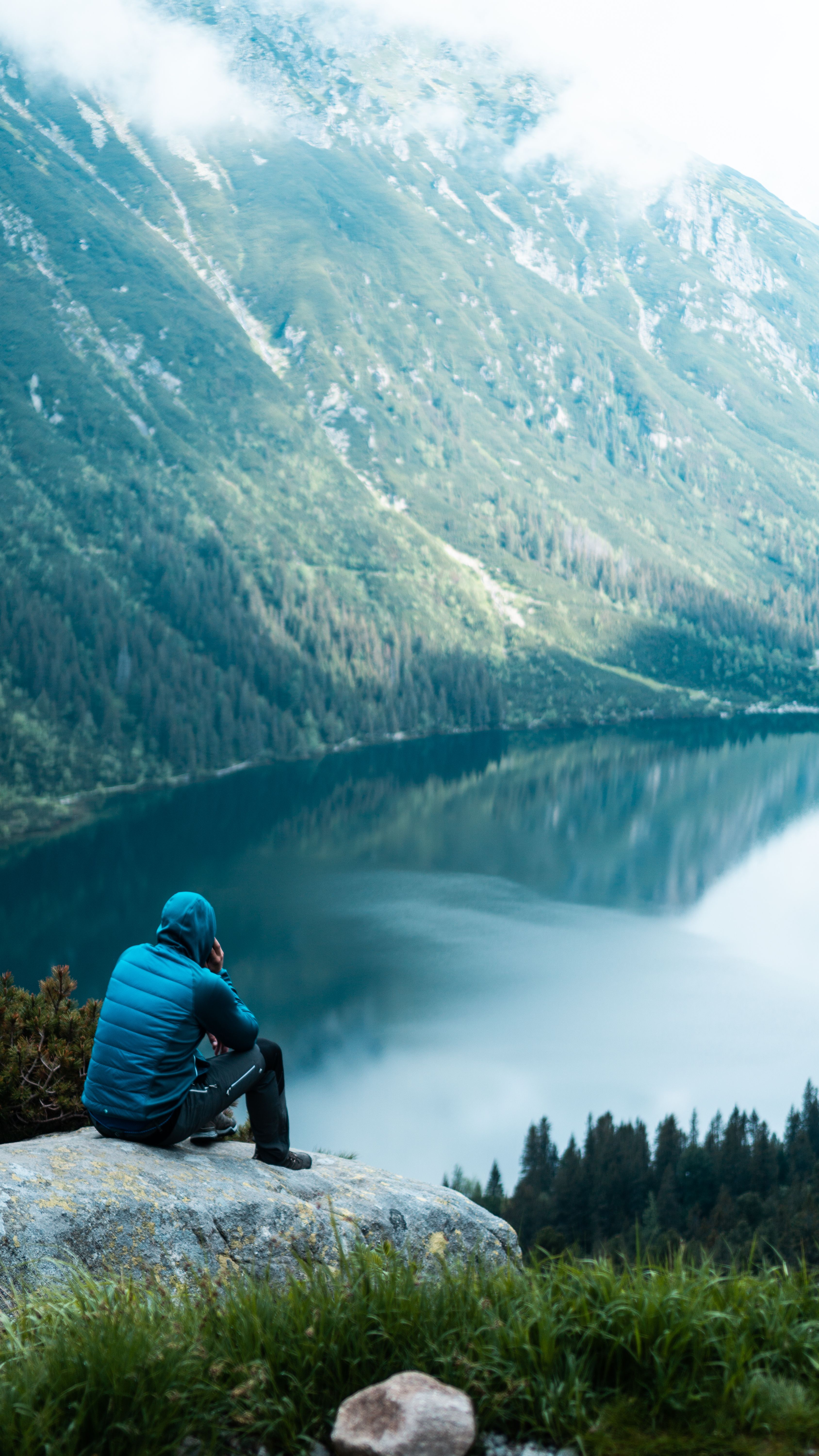 Person sits on a rock overlooking a tranquil mountain lake, surrounded by nature.