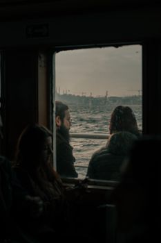 Two passengers on a ferry looking out at the ocean view through a cabin window.