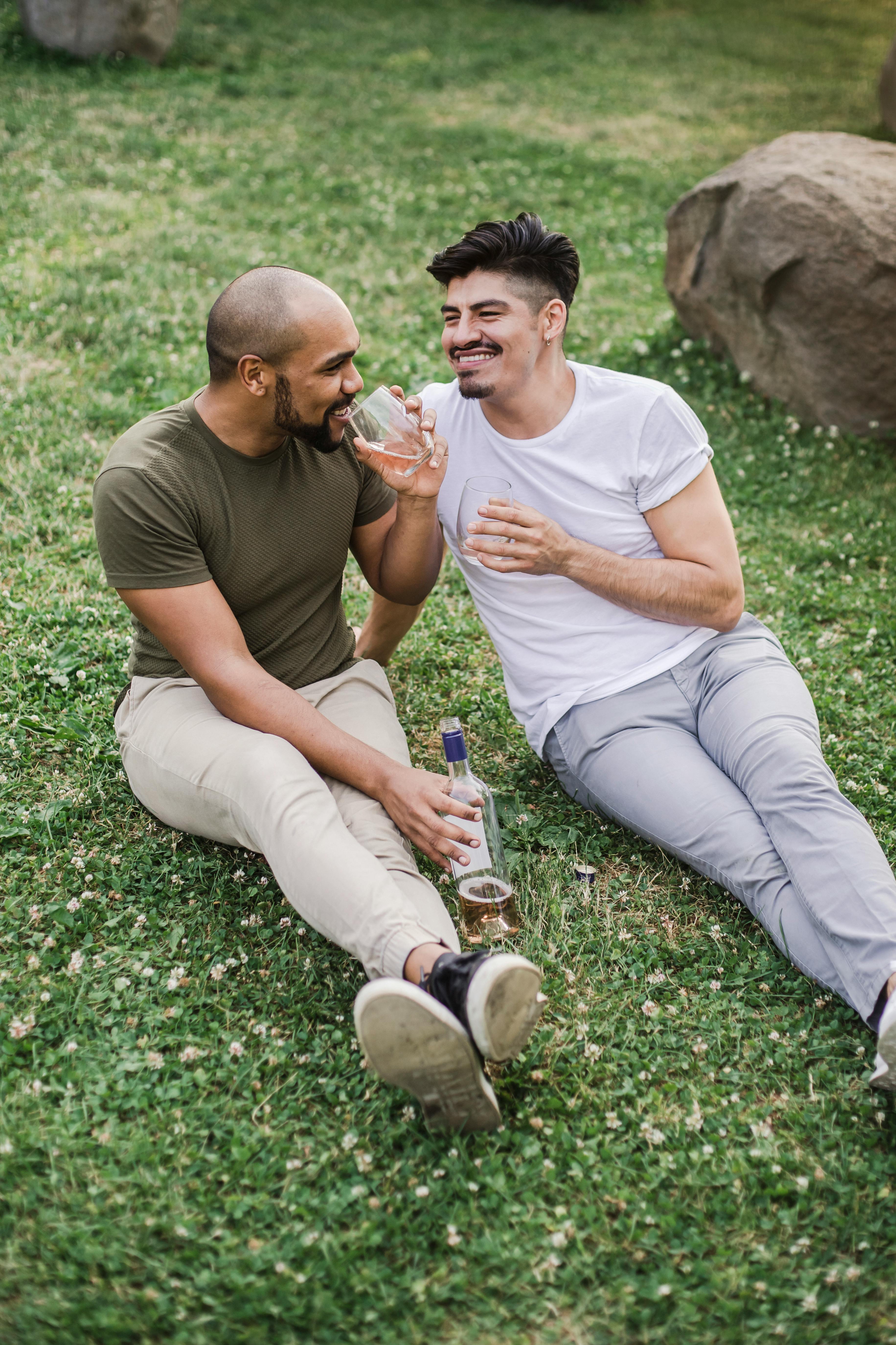 two happy men sitting on the grass and drinking wine