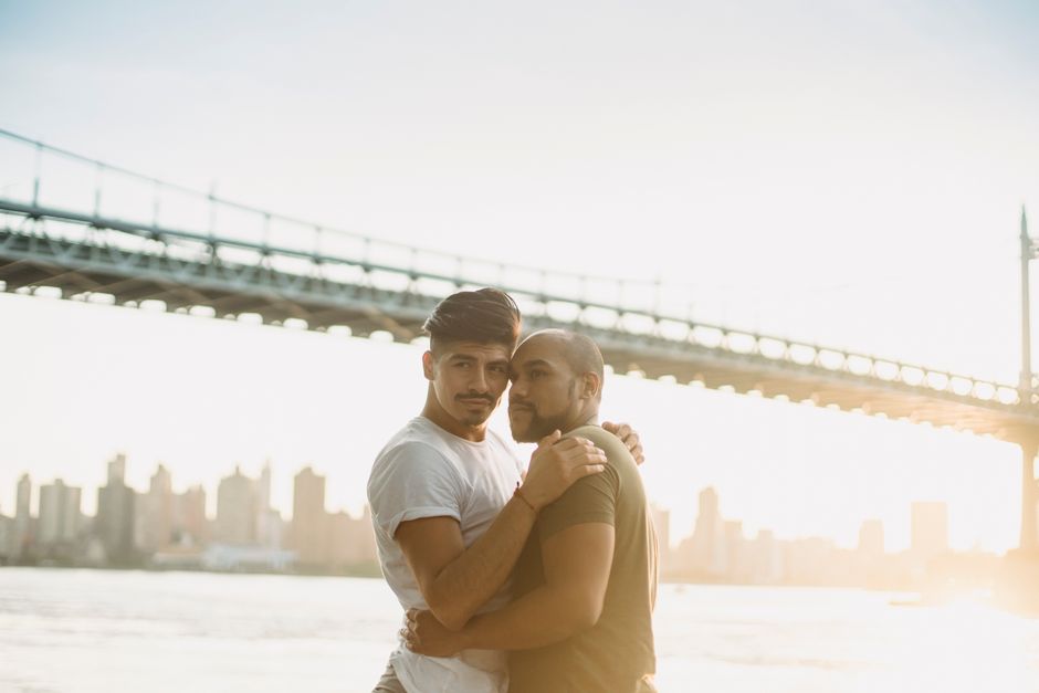 A loving gay couple embraces by the river with a city skyline and bridge in the background.