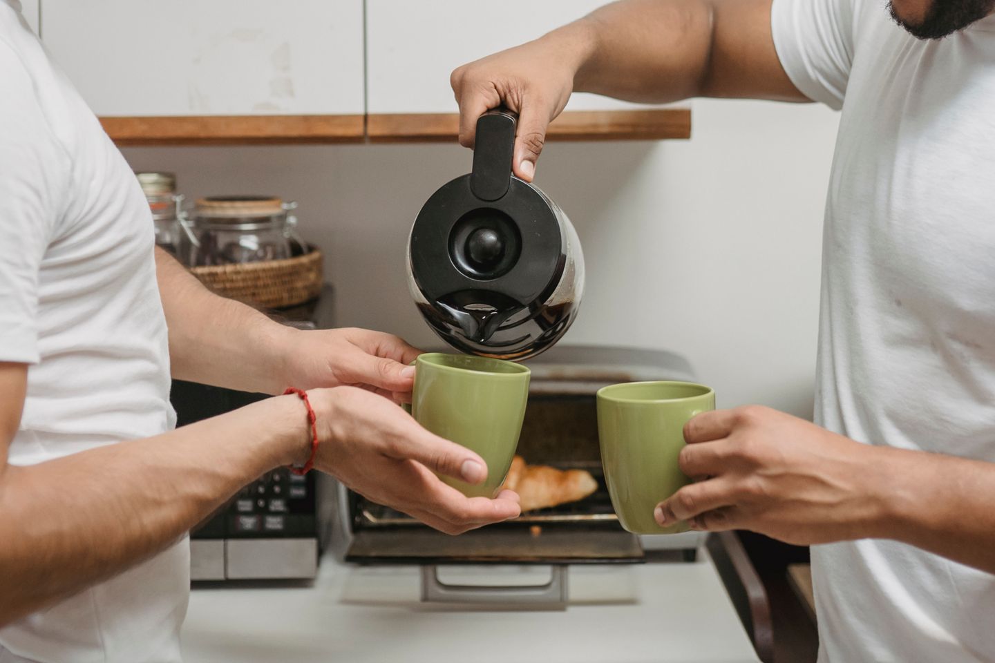 Several hands pour coffee into two green mugs at a kitchen counter.
