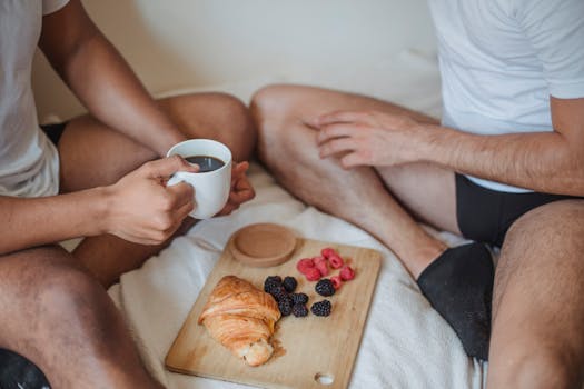 Intimate breakfast scene with croissant, coffee, and berries shared by two adults in a cozy bedroom setting.