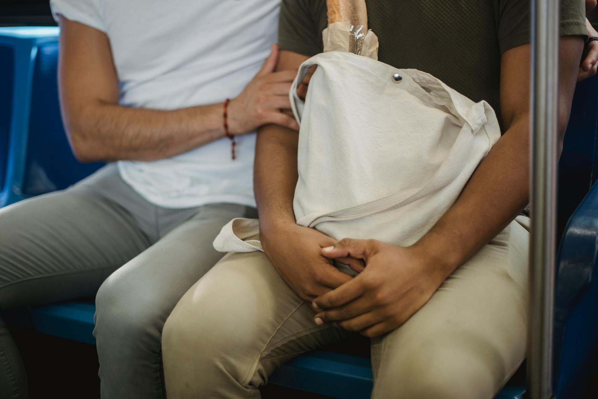 Two people sitting in a bus holding a canvas tote bag containing a baguette.