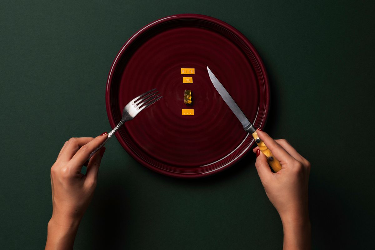 Artistic setup with fork and knife surrounding a gummy bear on a plate with words 'BITE ME.'