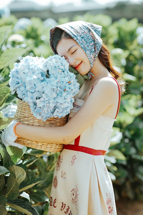 Free Side view of pleased Asian female gardener in headscarf standing with eyes closed near green bushes with flowers in wicker bucket in hands Stock Photo