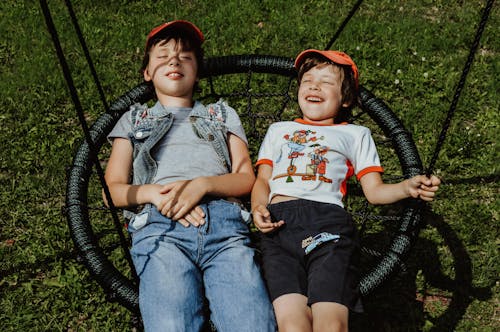 From above of happy boys with closed eyes wearing caps lying on round swing on grassy lawn while having fun in yard