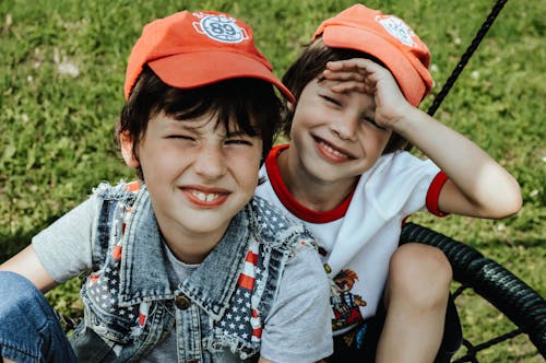 Free Smiling boys in caps looking at camera while sitting on round swing on green lawn in sunny park while having fun together Stock Photo