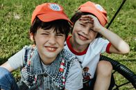 Smiling boys in caps looking at camera while sitting on round swing on green lawn in sunny park while having fun together