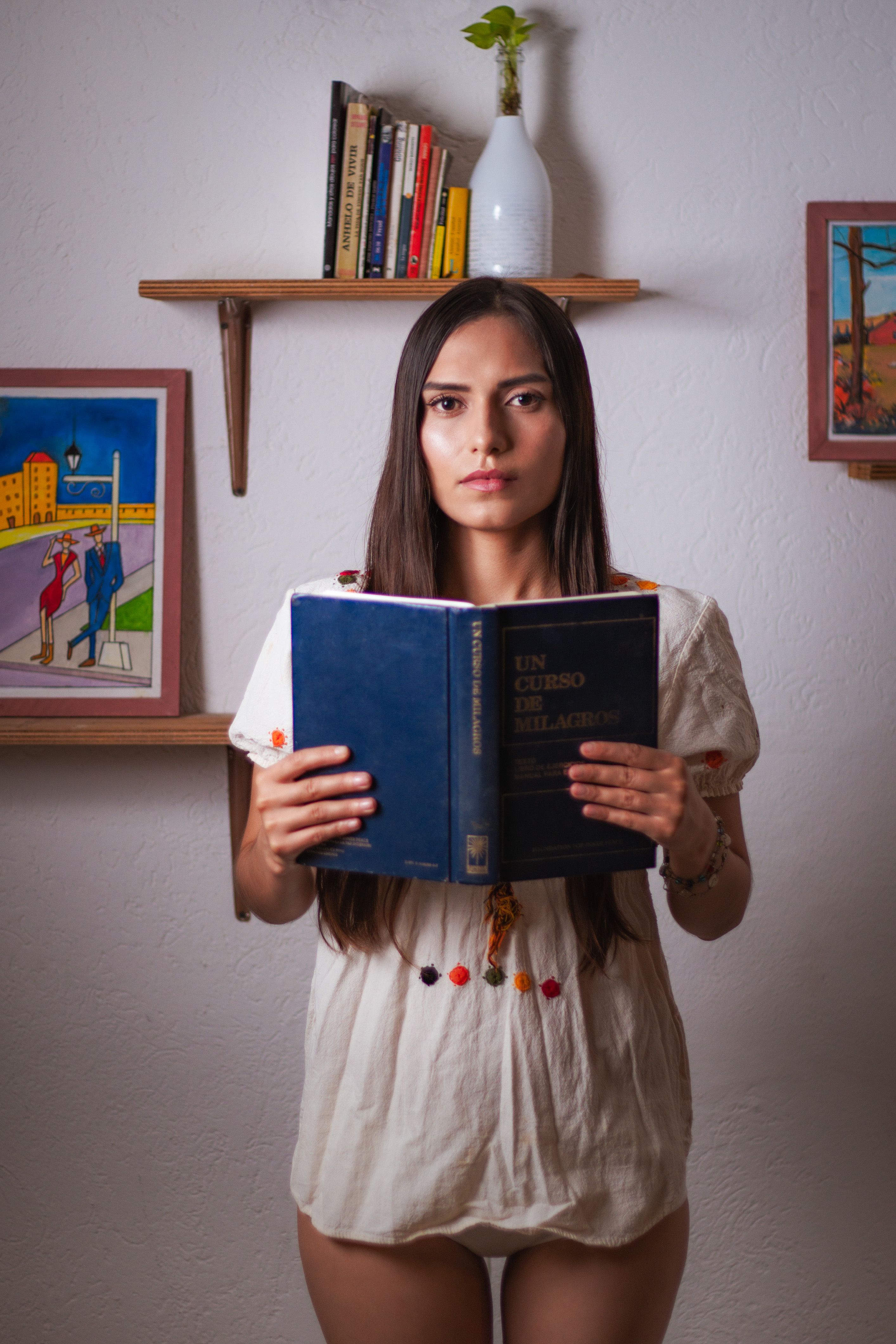 A young woman with a serious expression reads a book indoors, surrounded by art and shelves.