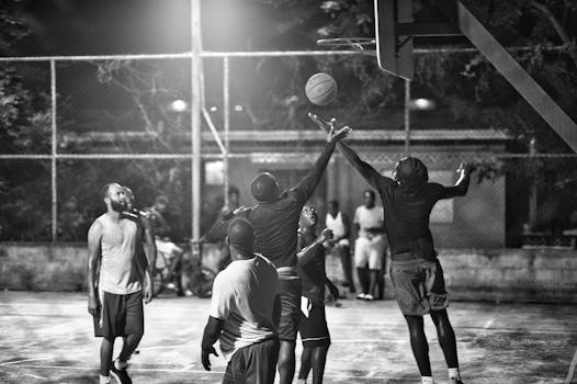 Black and white photo of men playing a basketball game at night on an outdoor court.