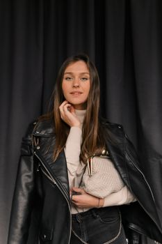 A fashionable woman poses indoors wearing a leather jacket and holding a textured bag.