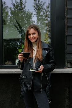 一位穿着黑色皮夹克的时尚年轻女子在户外拿着咖啡杯