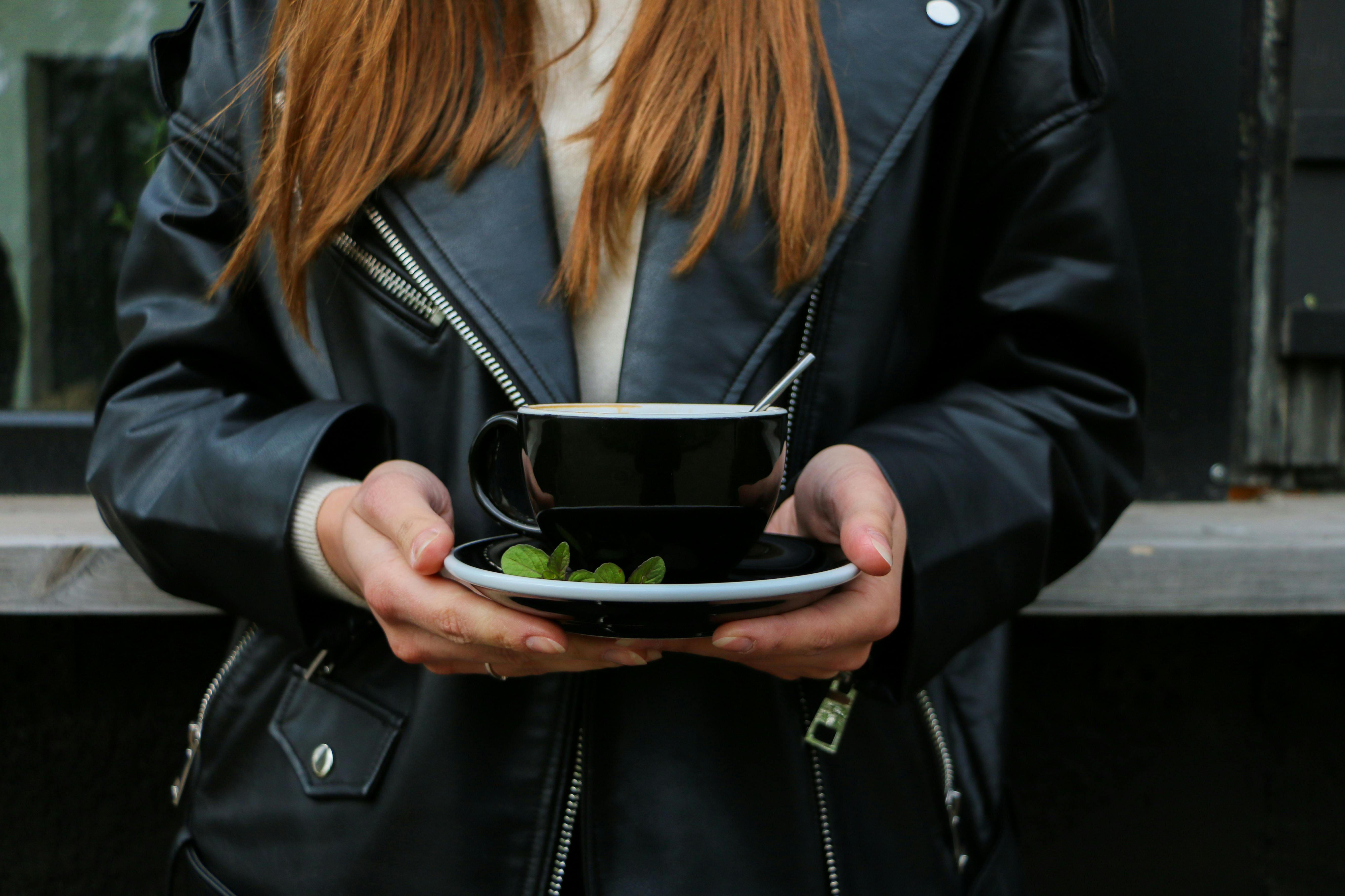 一名穿着皮夹克的女子手持一个涂有薄荷叶的黑色咖啡杯的特写镜头