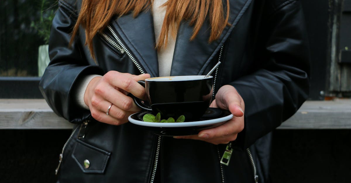 Close-up of a person in a leather jacket holding a black ceramic mug with mint leaves on the saucer.