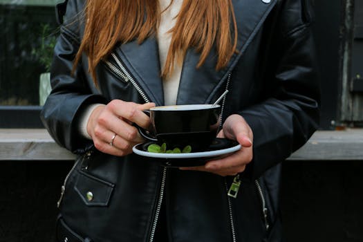 Close-up of a person in a leather jacket holding a black ceramic mug with mint leaves on the saucer.