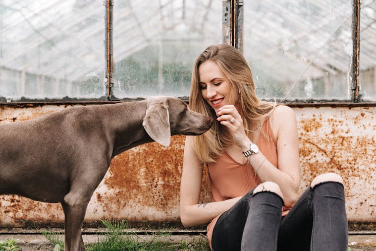 A woman sits against a rustic wall, enjoying a moment with her Weimaraner dog.