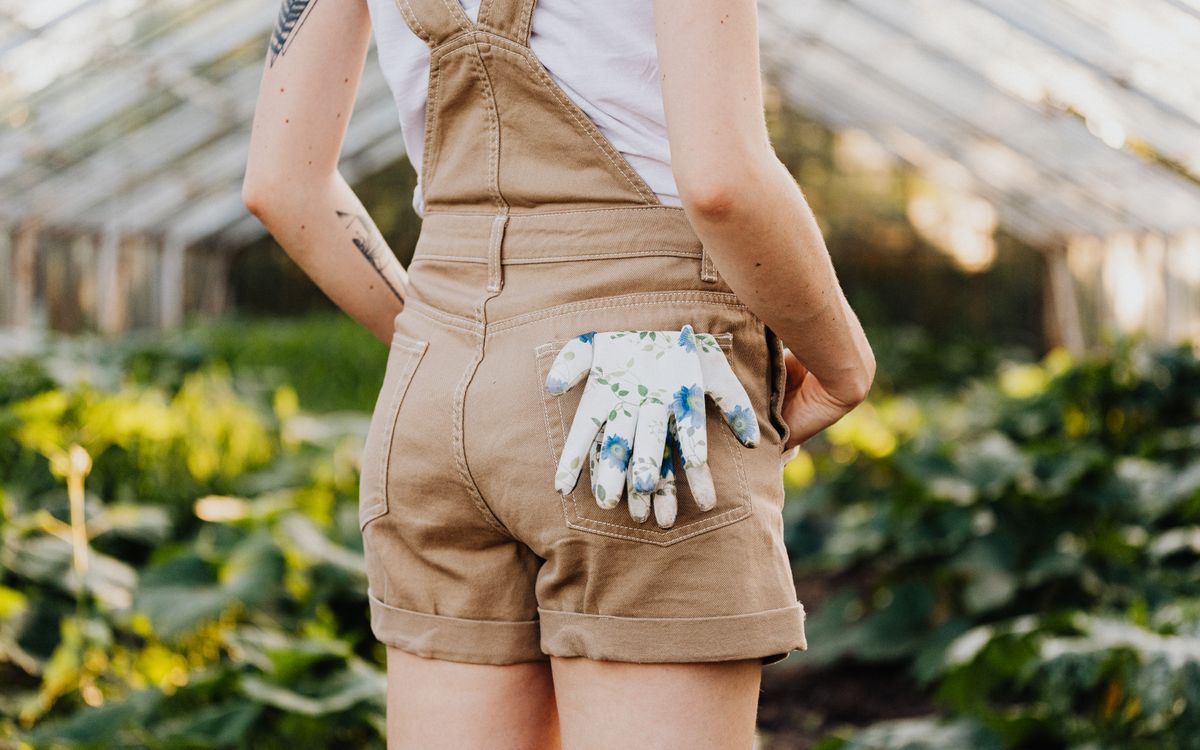 A woman in a garden wearing overalls with floral gloves in pocket, outdoor setting.
