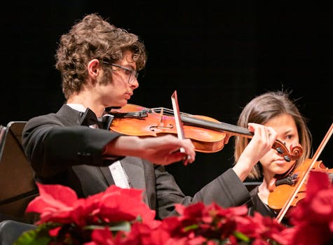 A young violinist in a black suit performs passionately during a concert surrounded by vivid red orchids.