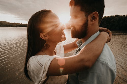 Side view of loving couple standing together near rippling lake and hugging while looking at each other with toothy smile in evening time