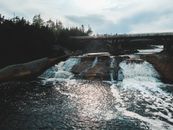 Spectacular view of shiny river with fast waterfalls under bridge near trees under cloudy sky
