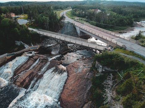 Aerial view of aged bridges over narrow river and cascades with rapid water fluids in mountains surrounded by green forest