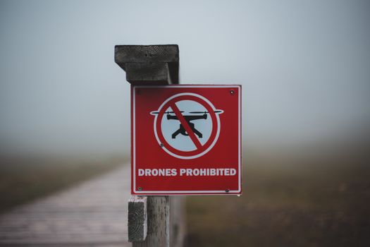 Sign prohibiting use of drones placed on wooden fence near wooden walkway in field in foggy day