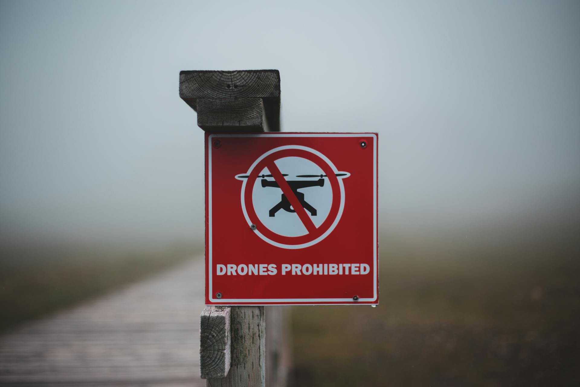 Sign prohibiting use of drones placed on wooden fence near wooden walkway in field in foggy day