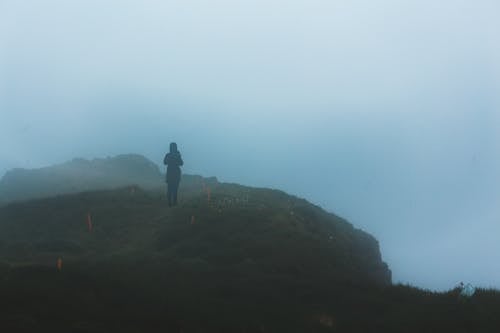 Anonymous person standing on grassy hillside in highlands in foggy weather at sunrise