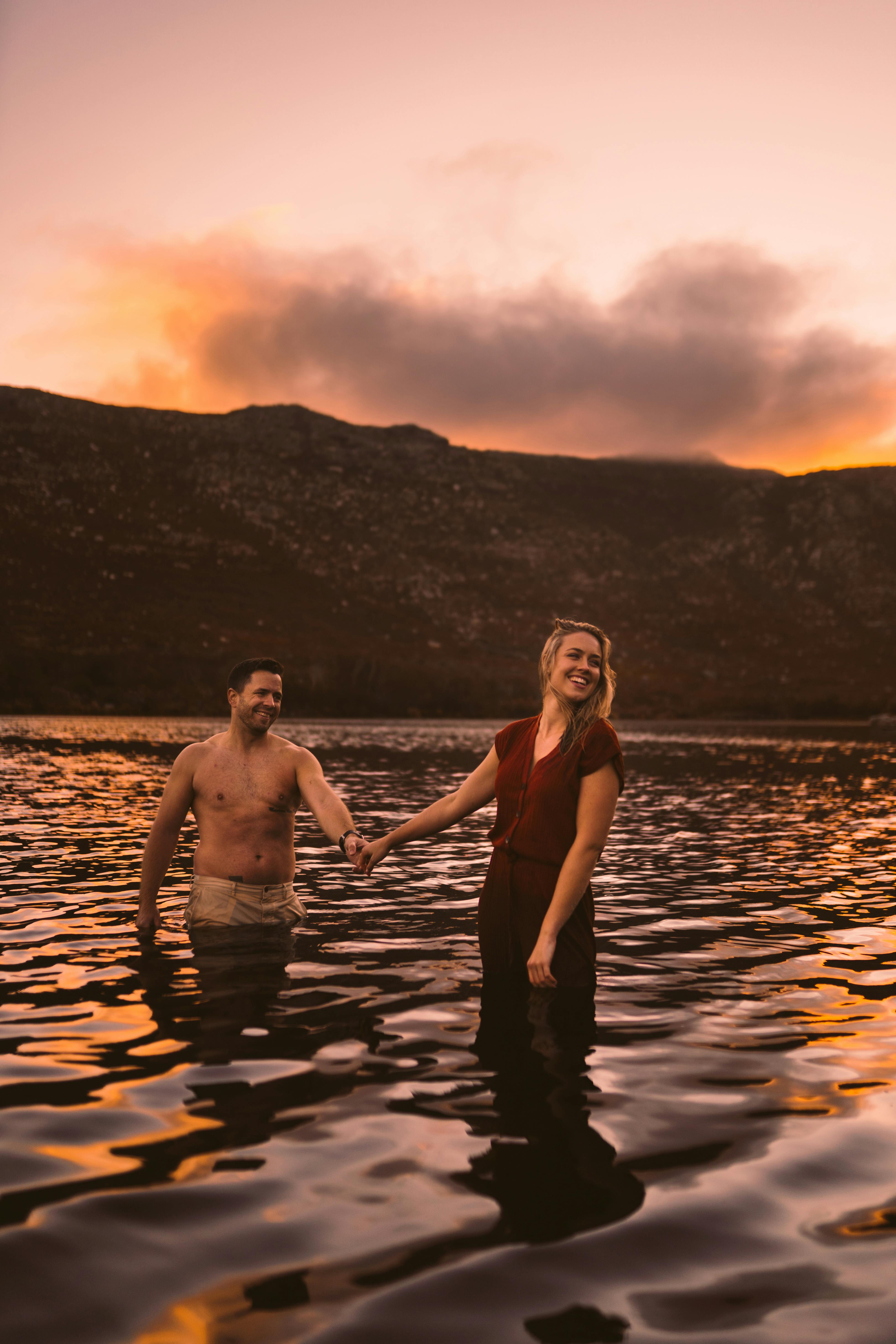 Happy couple holding hands in a serene lake at sunset, surrounded by mountains.