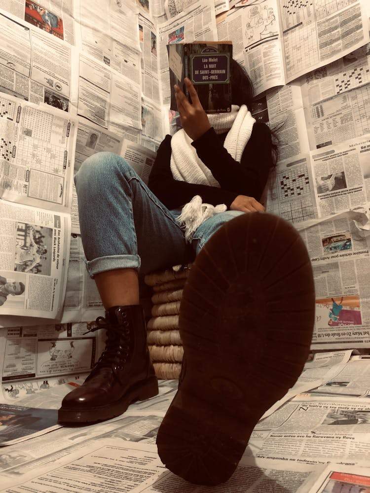 Woman Sitting In Corner Covered In Newspaper Pages Reading Book