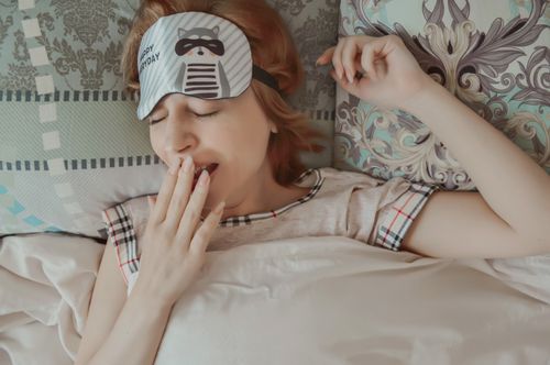 Free Top view of female with sleep mask wearing pajama covering mouth with hand while lying on bed with closed eyes under blanket during bedtime Stock Photo
