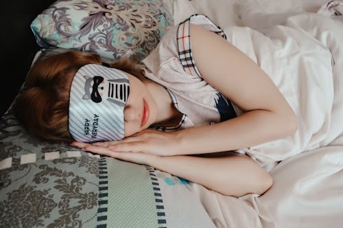 Anonymous female covering eyes with sleep mask taking nap while lying on bed under blanket in cozy bedroom at home