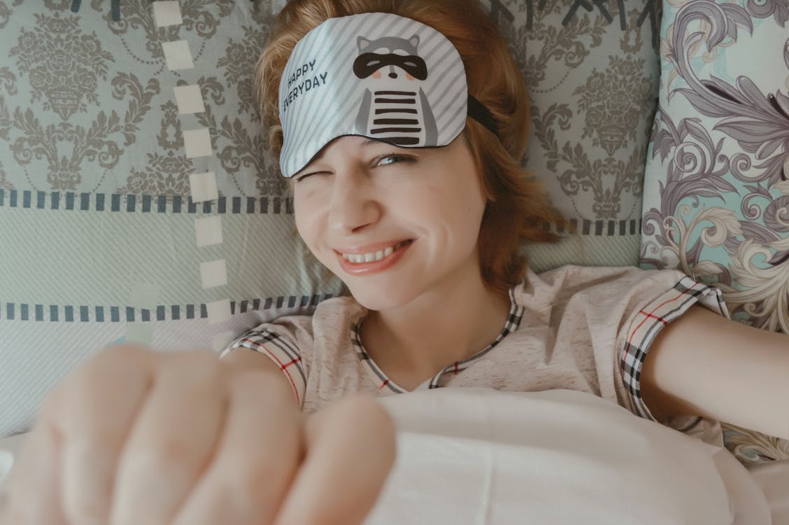 Free Top view of joyful female reaching out hands to camera and blinking while lying on bed under blanket and looking at camera Stock Photo