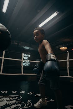A determined young boxer trains in an indoor boxing ring under dim lighting.