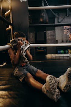 A young boxer sits in a boxing ring, holding a towel, after an intense workout indoors.