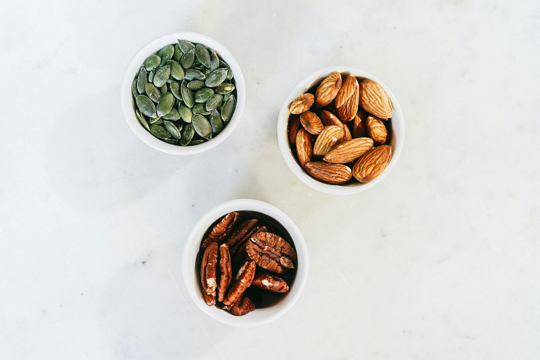 A top-down view of assorted nuts in bowls, featuring almonds, pumpkin seeds, and pecans, on a marble surface.