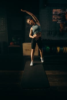 A woman performing a stretching exercise on a yoga mat in a dimly lit gym setting.