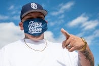 Ethnic man with tattoos in white t shirt and cap and protective mask with Jesus Saves inscription pointing finger at camera