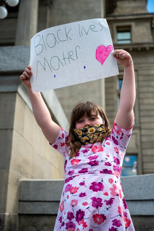 From below young girl in dress with colorful protecting mask standing on street and showing painted sign with Black Lives Matter inscription
