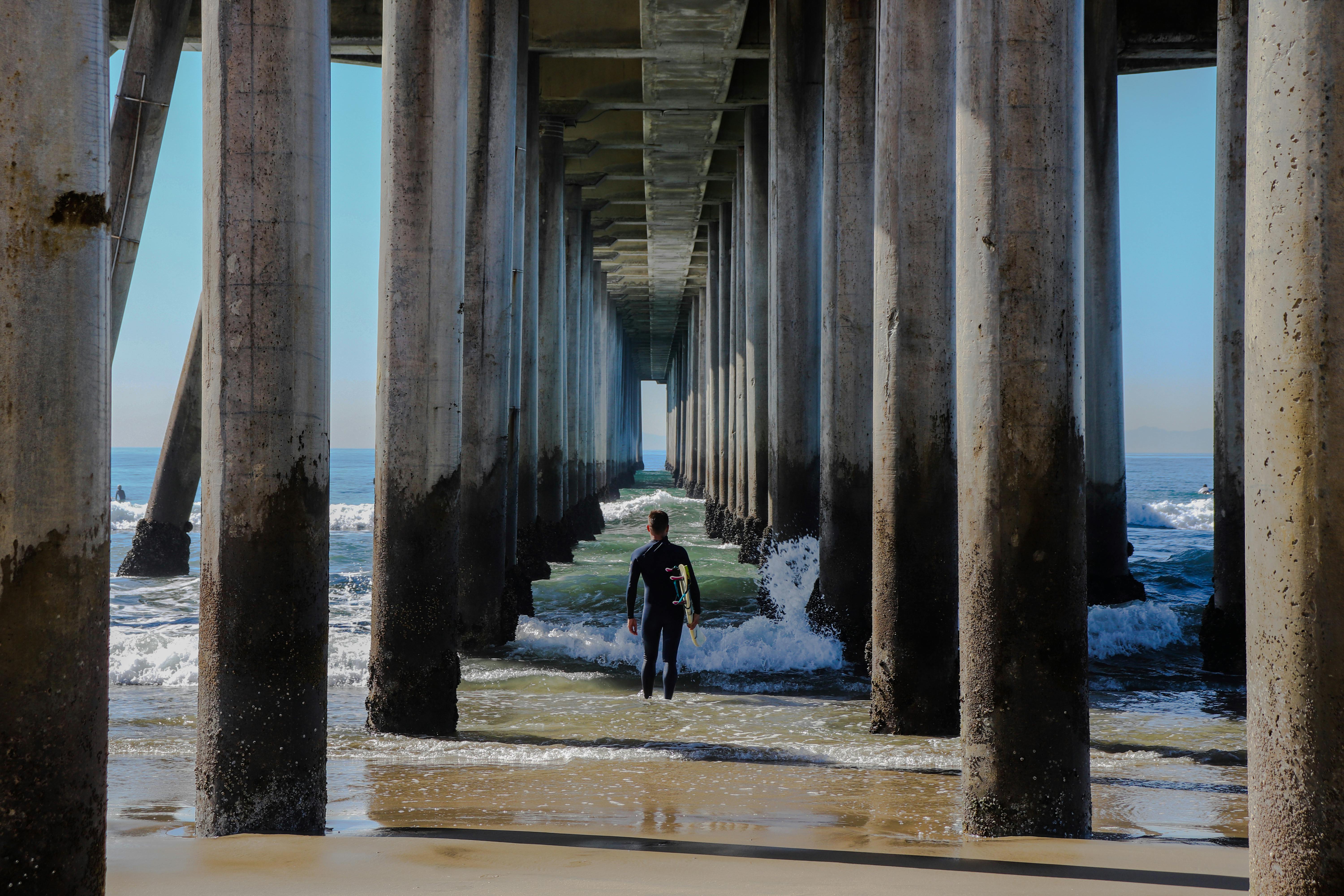 A surfer walks beneath the iconic Huntington Beach Pier, heading towards the ocean waves.
