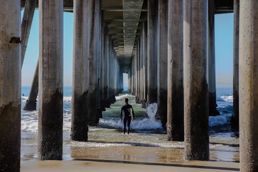 A surfer walks beneath the iconic Huntington Beach Pier, heading towards the ocean waves.