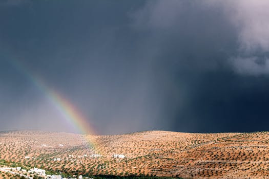 A vibrant rainbow arches over a rural landscape in Aqirbat, enhancing a dramatic sky.
