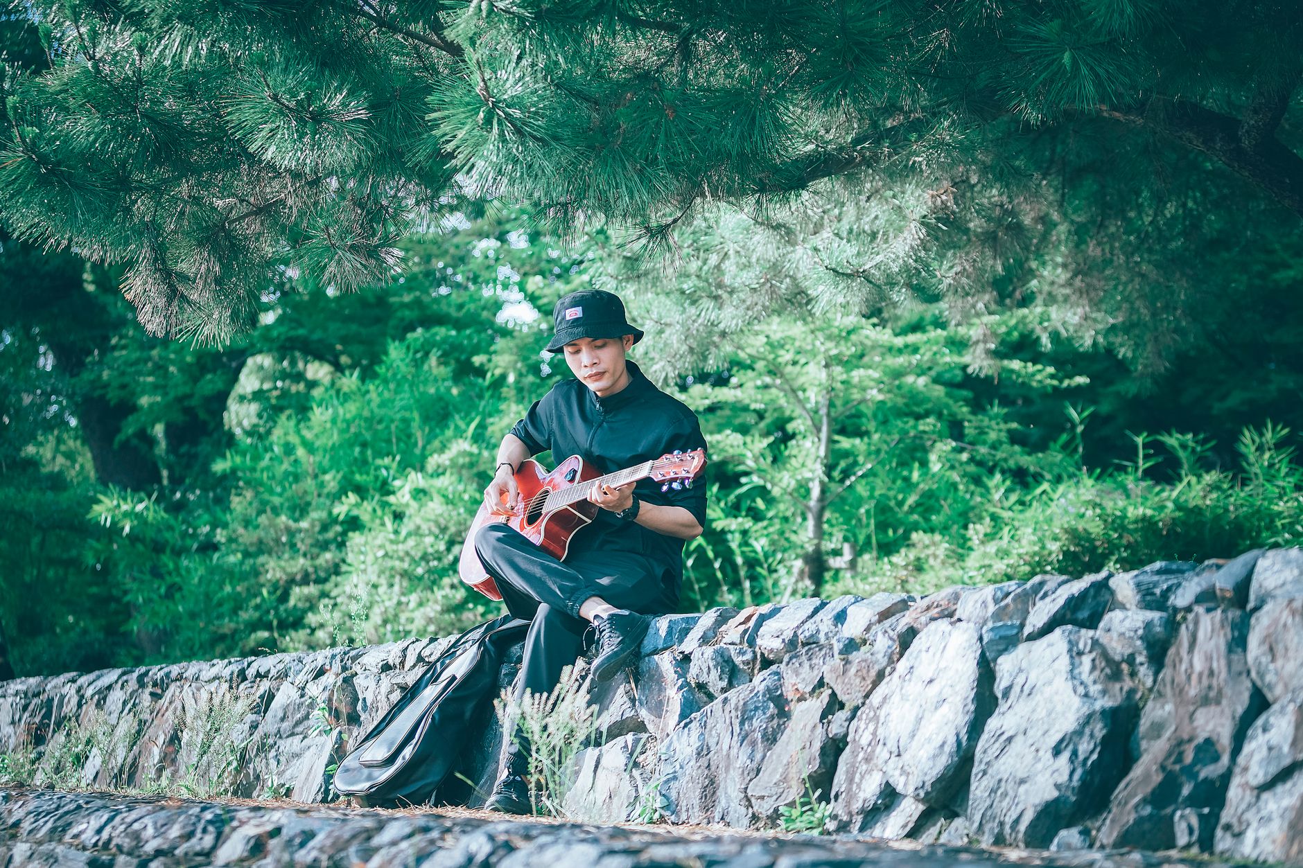 A young man sits outdoors playing guitar under lush trees, enjoying leisure time.