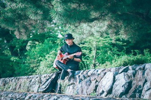 Free A young man sits outdoors playing guitar under lush trees, enjoying leisure time. Stock Photo
