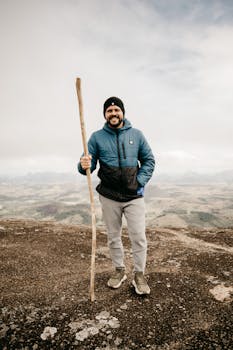 A cheerful man in winter clothes hiking on a scenic highland with a walking stick, enjoying the vast natural landscape.