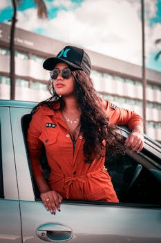 Young stylish ethnic female with long hair in sunglasses and fashionable outfit looking away in window of automobile on street