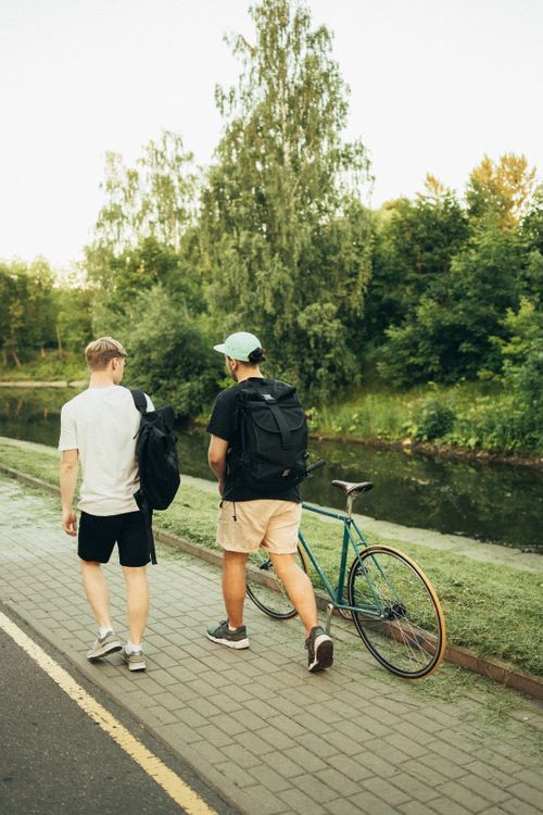 Free Two men with backpacks walking by a river with a bicycle on a sunny day. Stock Photo