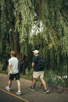 Two men with backpacks walking alongside a green bicycle under a willow tree on a park path.