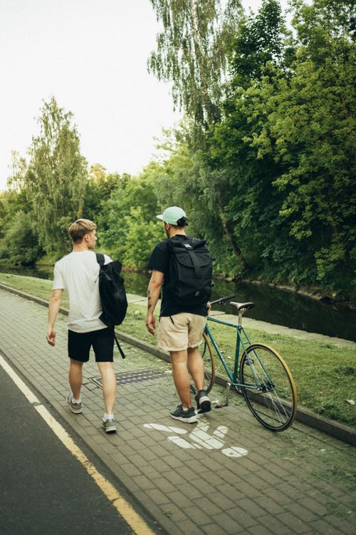 Free Two men walking on a sidewalk with a bicycle and backpacks in a green park setting. Stock Photo