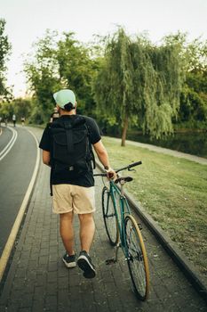 一名背着背包的男子走在公园绿树成荫的小路上，旁边跟着自行车